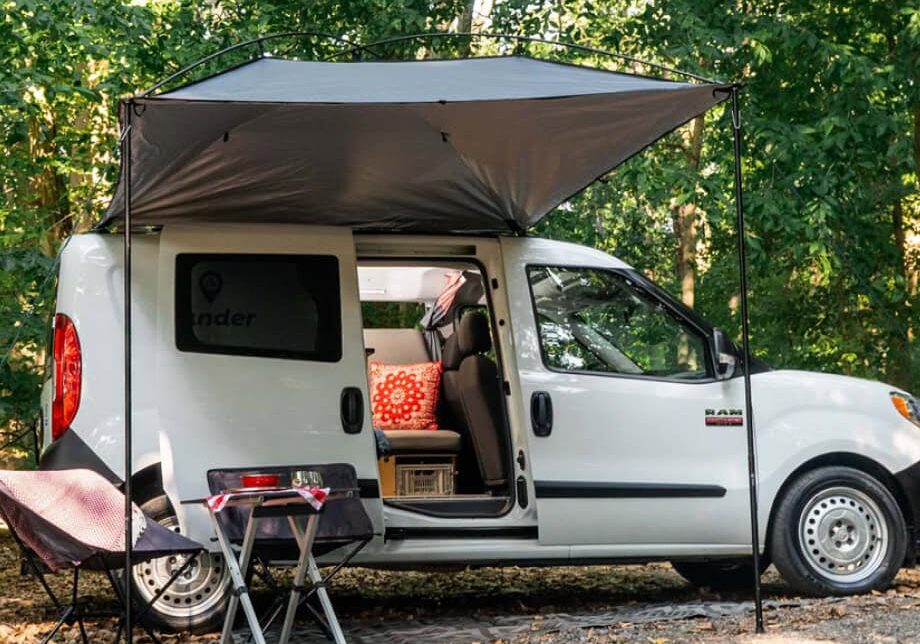 Small white campervan set up with an awning and camping chairs in a wooded campsite.