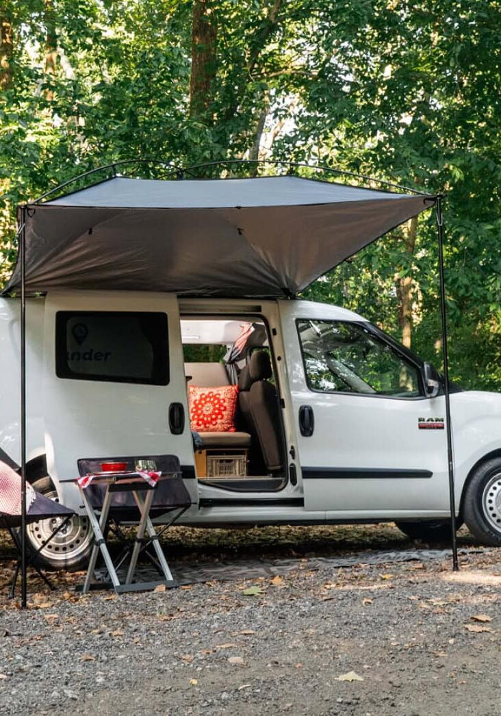 Small white campervan set up with an awning and camping chairs in a wooded campsite.